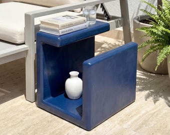 May include: A modern blue side table with a white vase inside. The table has a rectangular shape with a shelf on top holding books and glasses. A beige lounge chair with a pillow is in the background.