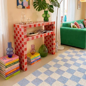 May include: A living room scene with a green corduroy sofa, a red and pink tiled console table, and a blue and white checkered rug. Artwork hangs on the peach-colored wall. Decorative vases and books add color.
