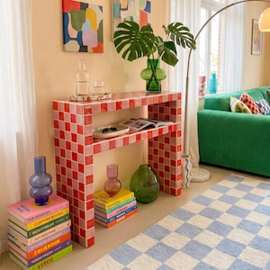 May include: A living room scene with a green corduroy sofa, a red and pink tiled console table, and a blue and white checkered rug. Artwork hangs on the peach-colored wall. Decorative vases and books add color.