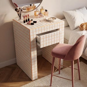 May include: A vanity table with a grid tile design, a round mirror, and a pink velvet stool. The table holds perfumes, lipsticks, and makeup brushes. A ginger cat rests on the bed in the background. The room has a neutral color palette.