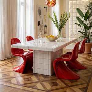 May include: A dining room featuring a white tiled table with red chairs. Above the table hang several spherical pendant lights in silver and copper finishes. The room has hardwood floors and large potted plants.