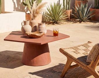 May include: A terracotta-colored hexagonal outdoor coffee table with a cylindrical base. The table is set with decorative vases, a wooden bowl, and a glass of iced tea. The scene is set in a sunny outdoor patio with cacti and a woven chair.
