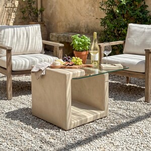 Può includere: Scena di patio all'aperto con due poltrone in legno con cuscini color crema, un tavolino beige con piano in vetro e un tagliere in legno con frutta e formaggio. Una bottiglia di vino e un bicchiere sono sul tavolo.