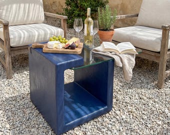 May include: Outdoor scene with wooden chairs, a blue side table, and a glass-topped table. A wooden cutting board holds cheese, grapes, and figs. A bottle of wine and glasses are on the table. A book and a potted plant are also present.