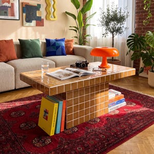 May include: A cozy living room scene with a beige sofa, colorful throw pillows, and a tiled coffee table. An orange lamp sits on the table, along with books and a camera. Artwork and plants decorate the walls, and a red rug covers the floor.