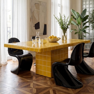 May include: A contemporary dining room with a yellow tiled table and black chairs. A distinctive light fitting is suspended above the table. The room includes large windows with sheer curtains, a painting, and potted plants.