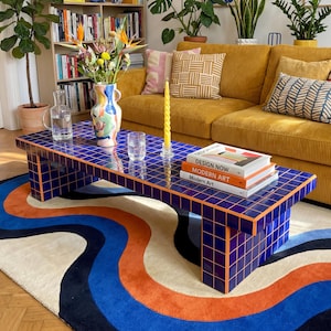 Könnte beinhalten: Ein Wohnzimmer mit einem senfgelben Cordsofa, einem blau gefliesten Couchtisch und einem bunten Teppich. Auf dem Couchtisch liegen Bücher und eine Vase mit Blumen. Pflanzen und Kunstwerke schmücken den Raum.