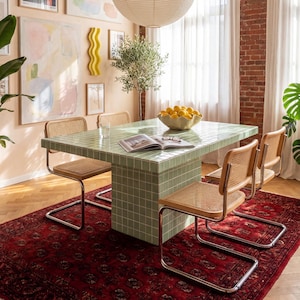 May include: A dining room scene featuring a square green tiled table with four chairs. The chairs have woven seats and chrome legs. A red patterned rug, artwork, and a large paper lantern complete the room's decor.