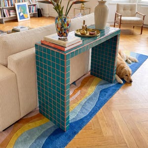 May include: A living room with a teal tiled console table, a lamp, and a vase of flowers. A beige corduroy sofa and a colourful rug with a wave pattern are also present. A golden retriever dog is resting on the rug.