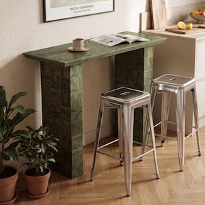 Op de afbeelding: Een rechthoekige consoletafel met een groene houtnerfafwerking en twee blokvormige poten, gecombineerd met twee zilverkleurige metalen krukken. Op de tafel staan een keramische mok en een open boek. Erboven hangt een ingelijste kunstdruk.