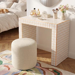 May include: A contemporary dressing table with a tiled grid pattern, a circular mirror, and a fluffy stool. The table displays perfumes, makeup brushes, and a vase of flowers. A cat is on a bed in the background.