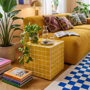 May include: A living room scene featuring a mustard yellow corduroy sofa with patterned throw pillows. A yellow tiled side table holds a glass carafe and glasses. A blue and white checkered rug and potted plants complete the decor.
