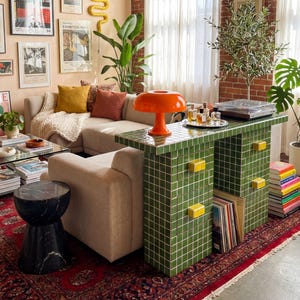 May include: A cozy living room with a beige sectional sofa, a green tiled console table, and a red patterned rug. The room is decorated with framed art, plants, and an orange mushroom lamp. A paper lantern hangs from the ceiling.