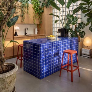 Könnte beinhalten: Moderne Küche mit blauer gefliester Insel und roten Hockern. Die Insel hat ein weißes Gittermuster. Holzschränke und üppige grüne Pflanzen umgeben den Raum. Eine schwarze Kaffeemaschine steht auf der Insel, und ein Oberlicht spendet natürliches Licht.