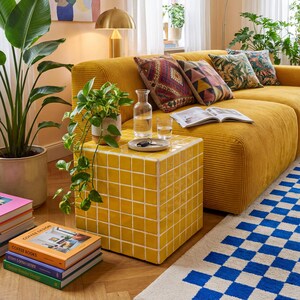 May include: A living room scene featuring a mustard yellow corduroy sofa with patterned throw pillows. A yellow tiled side table holds a glass carafe and glasses. A blue and white checkered rug and potted plants complete the decor.