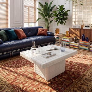 May include: A stylish living room featuring a blue leather sofa, a white tiled coffee table, and a patterned rug. The room is decorated with plants, books, and vases. Natural light enters from a skylight and windows.