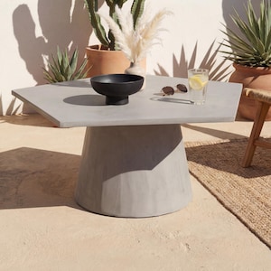 May include: A gray concrete outdoor coffee table with a black bowl, sunglasses, and a glass of water with lemon on top. The table is surrounded by potted plants, including cacti and succulents. The table is approximately 36 inches wide.