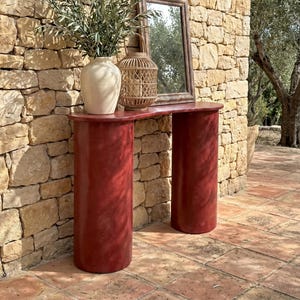 May include: A red console table with two cylindrical legs. A beige vase with green branches, a woven lantern, and a framed mirror sit on the table. The table is set against a stone wall.