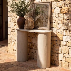 Può includere: Scena decorativa all'aperto con uno sfondo di muro in pietra. Un tavolino consolle semicircolare di colore chiaro sostiene un vaso marrone con vegetazione, una lanterna intrecciata e uno specchio incorniciato. La scena evoca un'estetica rustica e naturale.