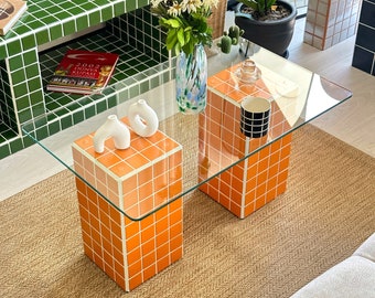 May include: Two orange and white tiled cube-shaped end tables with a glass top. The table is on a brown rug with a green tiled wall in the background.