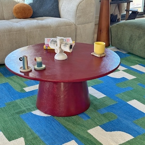 Könnte beinhalten: Ein runder, weinroter Couchtisch mit einem Sockel. Auf dem Tisch befinden sich kleine Keramikbecher, eine gelbe Tasse und ein weißer Projektor. Unter dem Tisch liegt ein farbenfroher Teppich mit blauen, grünen und weißen geometrischen Mustern.
