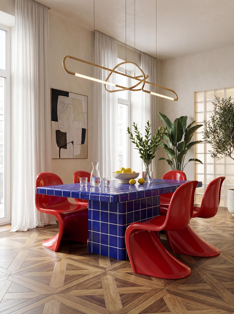 May include: A contemporary dining space with a blue tiled table and red chairs. A gold light fitting hangs above the table, and a vase of flowers and a bowl of lemons sit on the table. The room has wooden flooring and large windows.