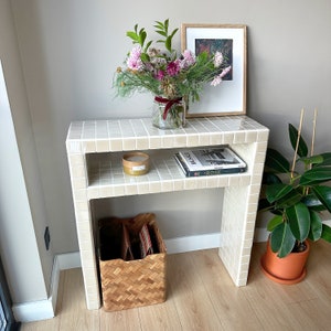 May include: A cream-tiled console table with a lower shelf, displaying a candle and books. A vase of pink and purple flowers and a framed picture adorn the top. A woven basket and a potted plant add to the room's decor.