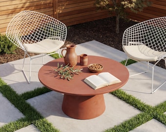 May include: Outdoor patio setting with a round, terracotta-colored table, two white wire chairs with cream cushions, and a wooden fence backdrop. The table holds a pitcher, small bowl, open book, and sprigs of greenery.