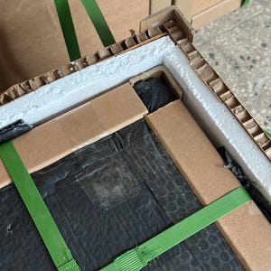 May include: A cardboard box secured with green straps, featuring foam padding and black plastic wrapping. The box is partially open, showing the contents. The packaging is designed to protect the item during transit.