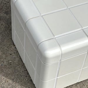 May include: A white, cube-shaped storage unit with a grid pattern on the top surface. The corners are rounded, and the unit appears to be made of a glossy material. The design is modern and minimalist.