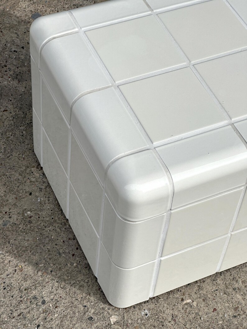 May include: A white, cube-shaped storage unit with a grid pattern on the top surface. The corners are rounded, and the unit appears to be made of a glossy material. The design is modern and minimalist.