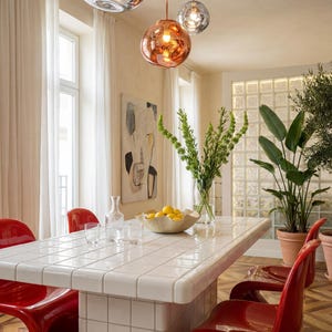 May include: A dining room featuring a white tiled table, red chairs, and a cluster of hanging globe lights. A vase of greenery and a bowl of lemons are on the table. Large potted plants and a painting are also present.