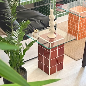 May include: A glass-topped side table with a red tiled base. The table is decorated with a white ceramic vase with a textured design and a small bouquet of flowers.