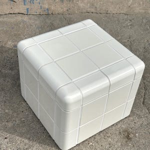 May include: A white, cube-shaped ottoman or small table. The surface is divided into a grid pattern, with rounded corners. The piece is made of a solid, opaque material and sits on a concrete surface.