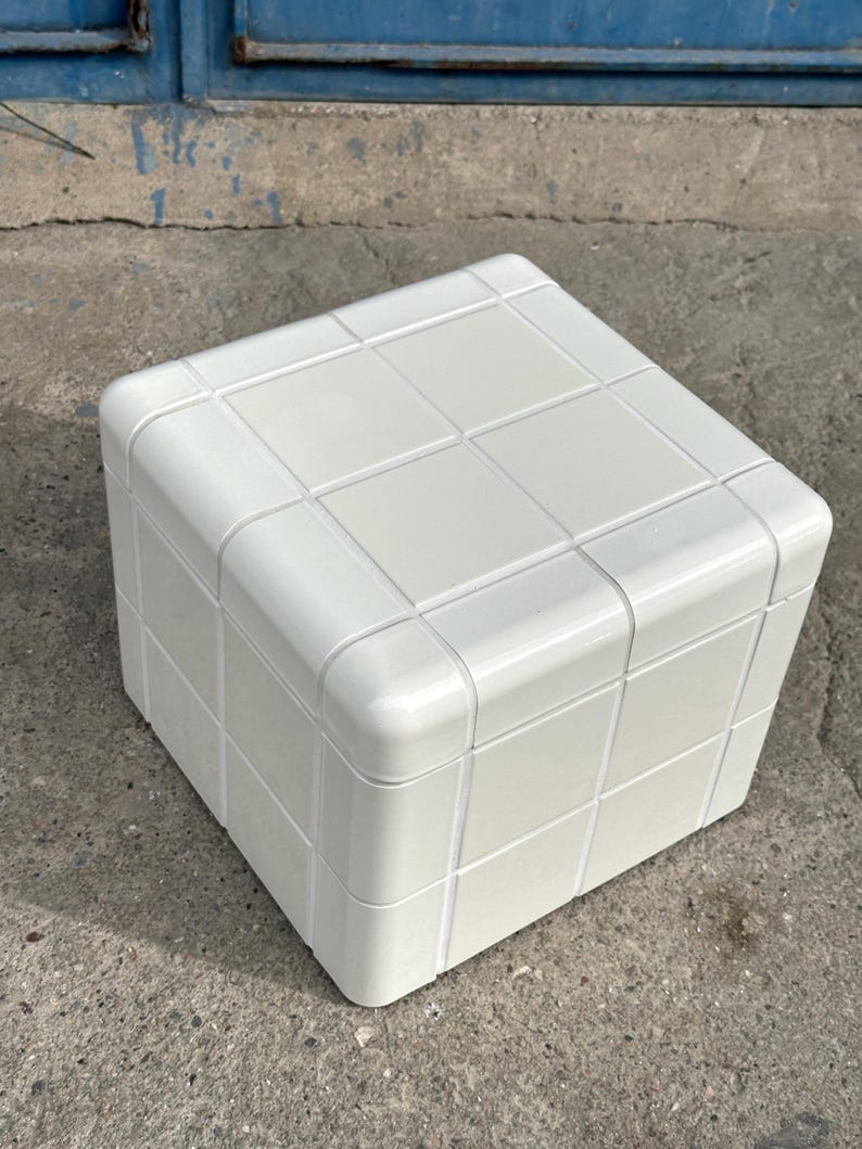 May include: A white, cube-shaped ottoman or small table. The surface is divided into a grid pattern, with rounded corners. The piece is made of a solid, opaque material and sits on a concrete surface.