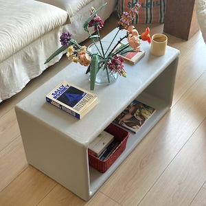 May include: A light gray, rectangular coffee table with a lower shelf. The table holds a vase of flowers, a book titled "Intermezzo", and a small red basket. A beige sofa is visible in the background.