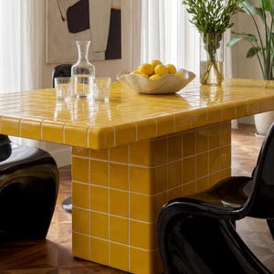 May include: A contemporary dining table with a vibrant yellow tiled surface. The table has a rectangular top and a single pedestal base. A bowl of lemons, a glass carafe, and two glasses are on the table. Black chairs surround the table, and a vase of greenery is visible.