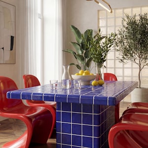 May include: A dining room with a blue tiled table and red chairs. The table is rectangular and has a grid pattern. A vase of lemons and a glass pitcher are on the table. A modern gold light fixture hangs above the table.