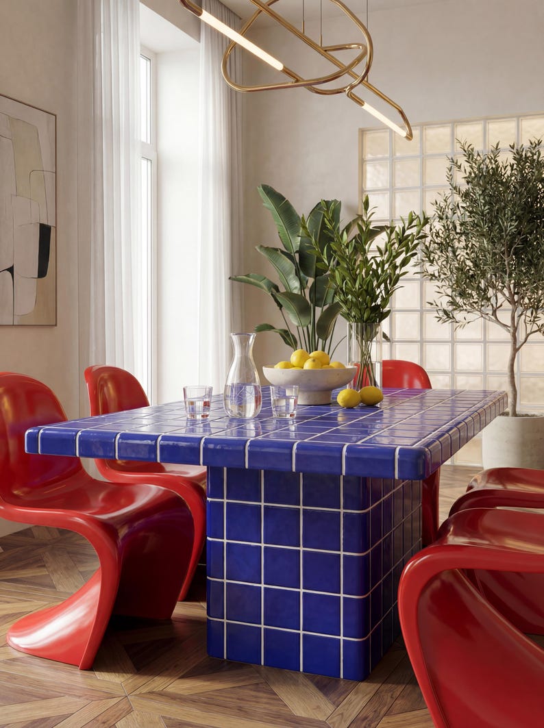 May include: A dining room with a blue tiled table and red chairs. The table is rectangular and has a grid pattern. A vase of lemons and a glass pitcher are on the table. A modern gold light fixture hangs above the table.