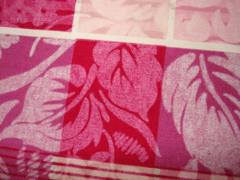 May include: Close-up of a fabric with a repeating pattern of squares. The squares feature a floral design in shades of pink and red. The fabric has a textured appearance, with a white grid pattern separating the squares.