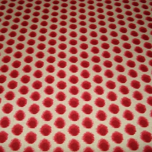 May include: Close-up of a fabric with a repeating pattern of red polka dots on a cream background. The dots are evenly spaced and have a slightly irregular, textured appearance, creating a playful and visually appealing design.