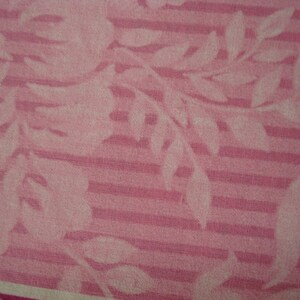May include: Pink fabric with a floral and striped pattern. White floral and leaf silhouettes are set against a pink striped background. The fabric has a soft, textured appearance.