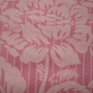 May include: Close-up of pink fabric with a floral design. The pattern shows large, stylised flowers and leaves in a lighter pink shade, set against a vertically striped background. The fabric has a textured appearance.