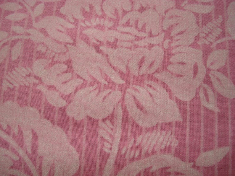 May include: Close-up of pink fabric with a floral design. The pattern shows large, stylised flowers and leaves in a lighter pink shade, set against a vertically striped background. The fabric has a textured appearance.