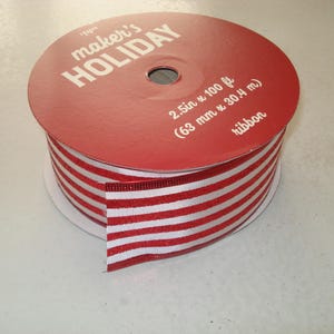 May include: A spool of red and white striped ribbon with a red cardboard label on top that reads maker's HOLIDAY, 2.5in x 100 ft, and ribbon. The ribbon features alternating horizontal red and white stripes and is wound around a central core.