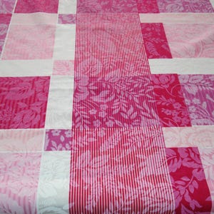 May include: A patchwork quilt featuring a variety of pink and white floral and striped patterns. The quilt is composed of rectangular blocks, with varying shades of pink and white, creating a visually appealing design. The floral patterns include roses and other botanical designs.