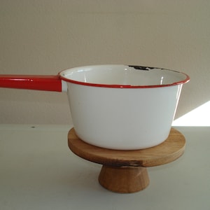 May include: A white enamel saucepan with a red handle and rim, resting on a small wooden pedestal. The saucepan has a classic design, perfect for cooking. The handle is a bright red, contrasting with the white body.