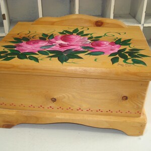 May include: A wooden box with a hinged lid painted with pink roses and green leaves. The box has a red trim around the bottom edge and the words "Jane Pratt" are painted on the top.