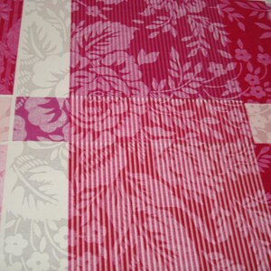 May include: Close-up of a patchwork quilt with a floral pattern. The quilt features squares of varying pink shades, including fuchsia, light pink, and white. Some squares have vertical stripes, while others display floral and leaf designs.