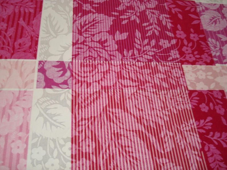 May include: Close-up of a patchwork quilt with a floral pattern. The quilt features squares of varying pink shades, including fuchsia, light pink, and white. Some squares have vertical stripes, while others display floral and leaf designs.